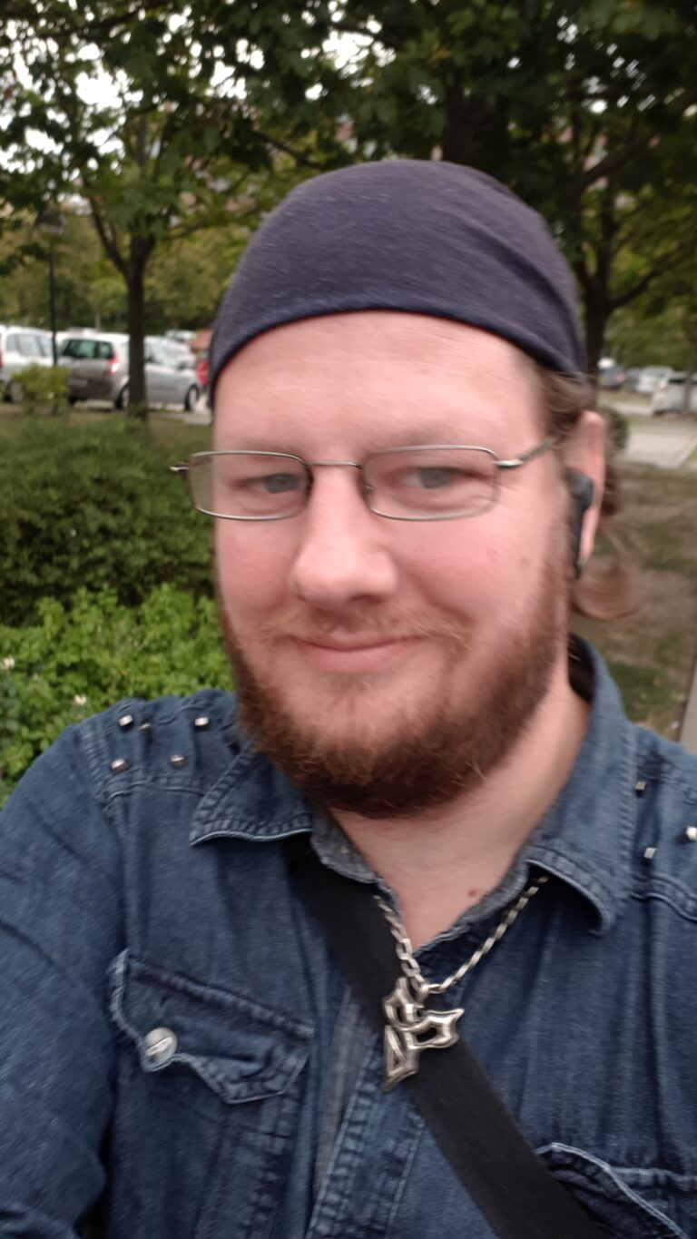 Mann mit Brille und Bart, schaut in die Kamera, freundliches Lächeln, im Hintergrund eine Parklandschaft